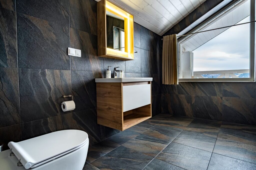 Interior of modern stylish bathroom with black tiled walls, curtain shower place and wooden furniture with wash basin and big illuminated mirror.