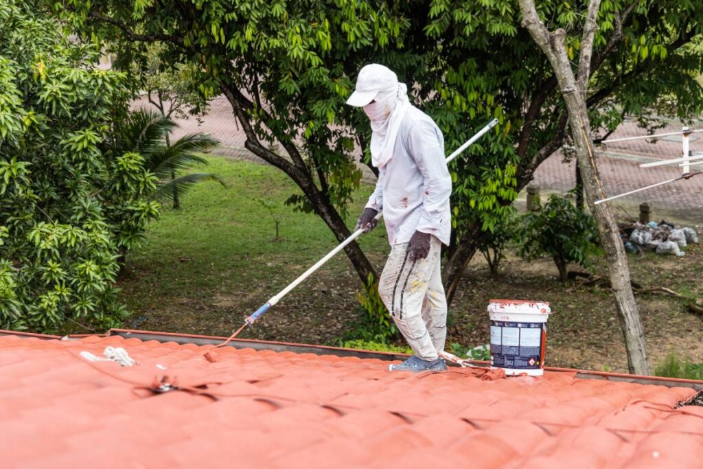 Worker painting red rooftop with roller at residential building after applying coat of foundation druing renovation